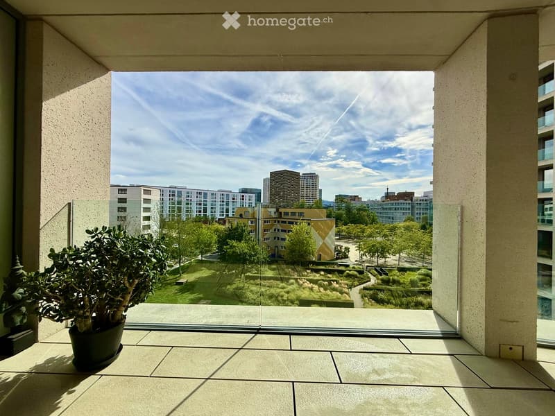 Balcony with tiled floor, glass railing, and a potted plant, overlooking a landscaped area with trees and buildings.