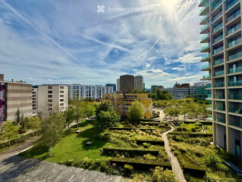 A landscaped garden area with green grass, trees, shrubs, and pathways, surrounded by modern apartment buildings.