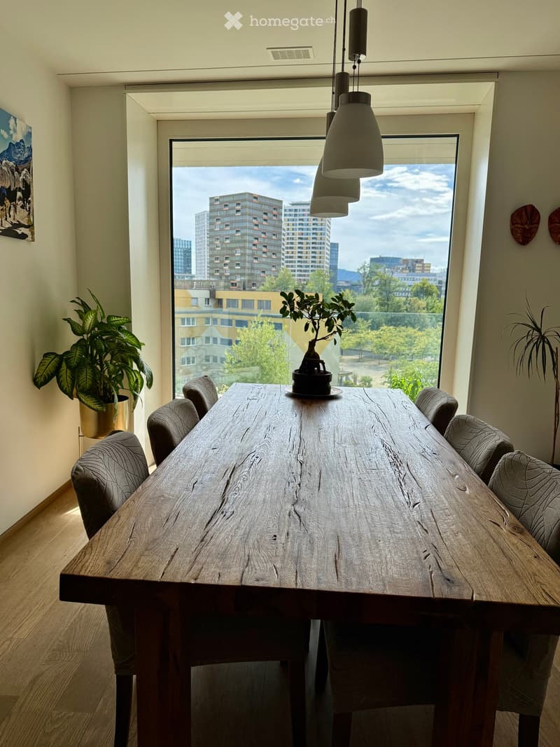 A modern dining area with a large wooden dining table surrounded by upholstered chairs, a potted plant, and a small tree on the table, with large windows offering a cityscape view.