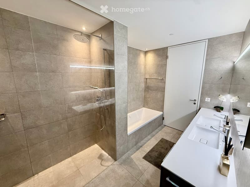 Modern bathroom with a bathtub, double sink, and a walk-in shower with a glass door. The space features beige tiles, a sleek white countertop, and decorative flowers on the sink.