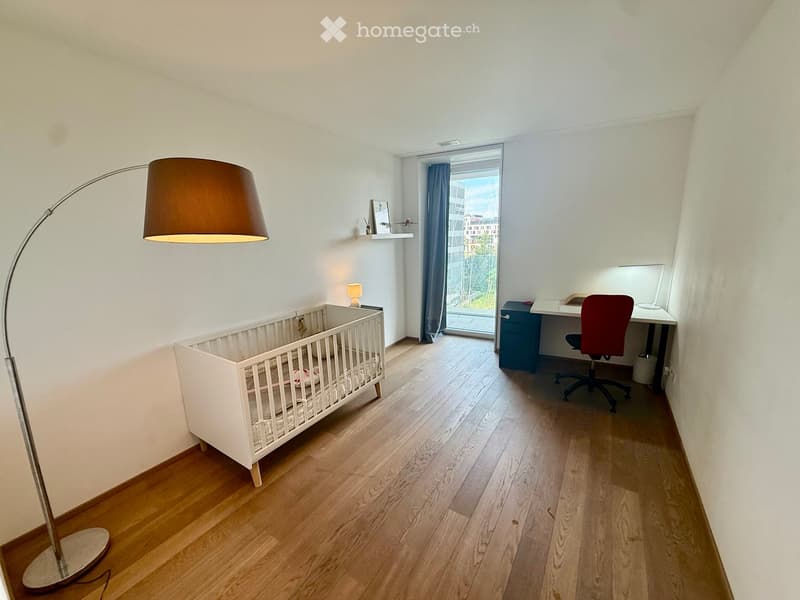 Bright room with wooden flooring, a baby crib, a desk with a chair, a floor lamp, and a small shelf with decorative items.
