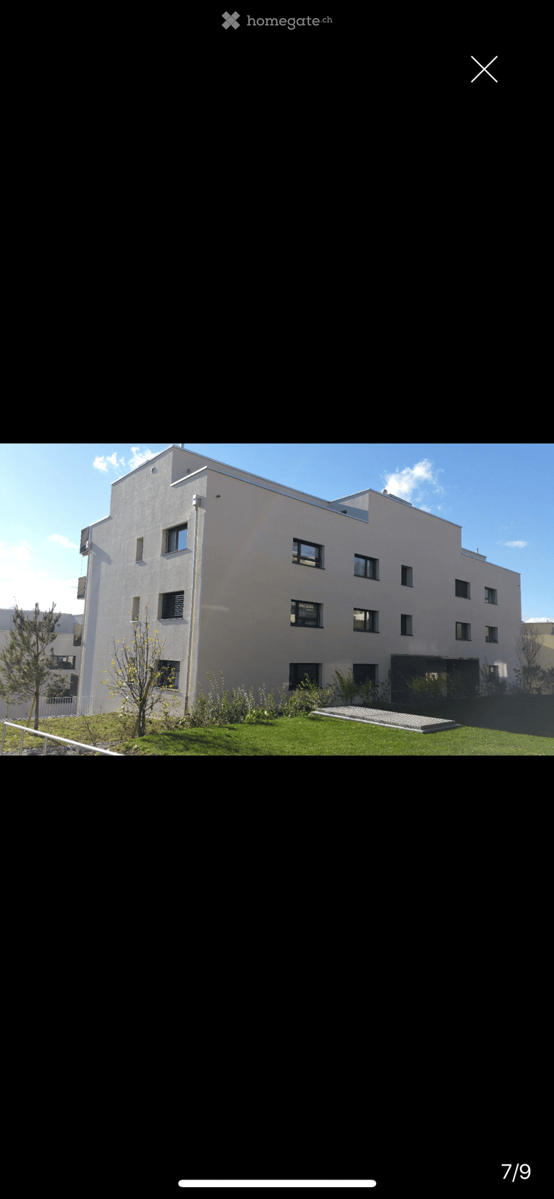 Modern multi-story building with white facade, multiple windows, and a small landscaped area in front.