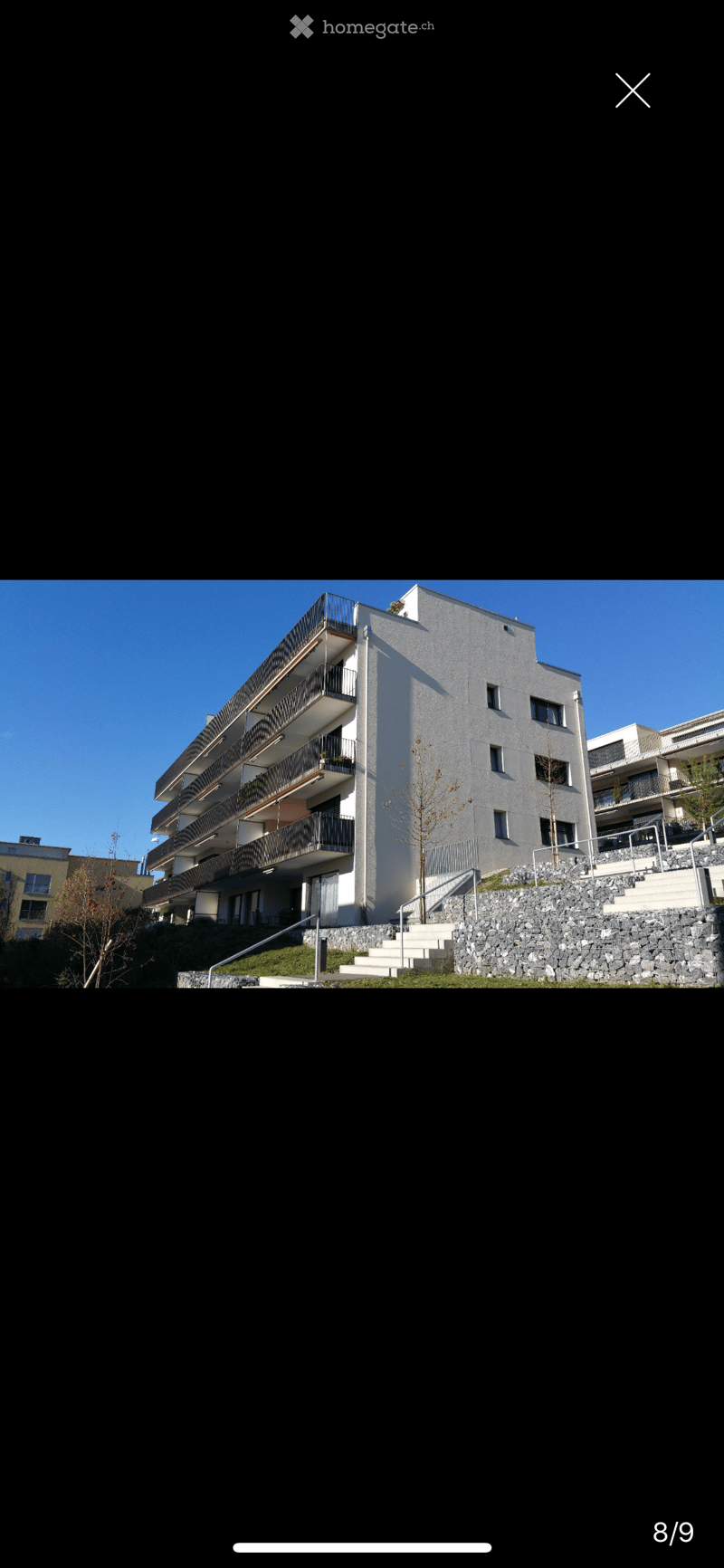 Modern multi-story residential building with balconies, raised ground floor, and stone staircase leading up to the entrance.