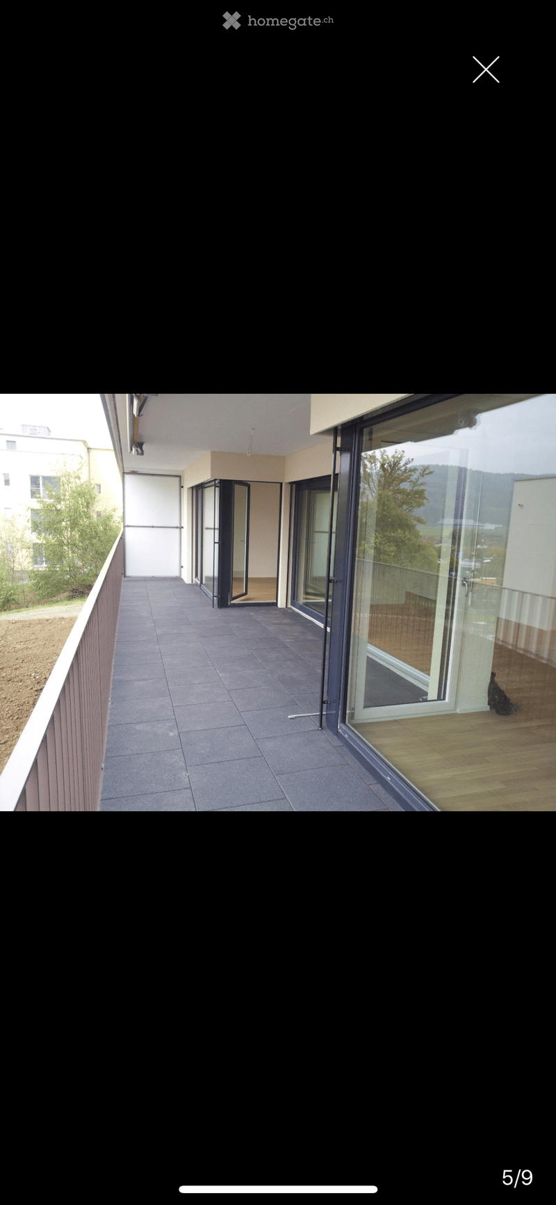 Modern balcony with grey tiled floor, glass sliding doors, wooden decking visible through the doors, and a view of greenery and buildings.