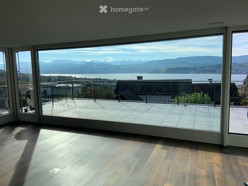 Spacious balcony with large glass doors leading to a tiled terrace, overlooking a scenic lake and mountain view.