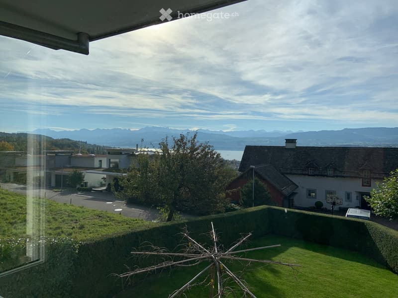 Balcony with a scenic view of mountains, a lake, and surrounding houses. The foreground includes a well-maintained lawn and a small decorative structure.