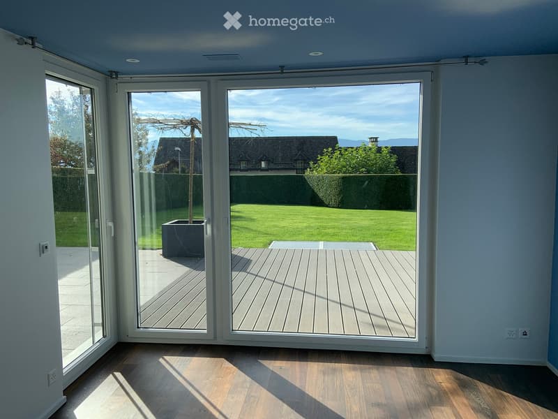 Bright, unfurnished room with large sliding glass doors opening to a wooden deck and a well-maintained lawn. The room features light wooden flooring and white walls.