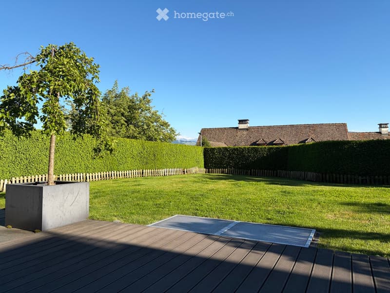 Well-maintained garden with a wooden deck, a tree in a planter, neatly trimmed hedges, and a house in the background.
