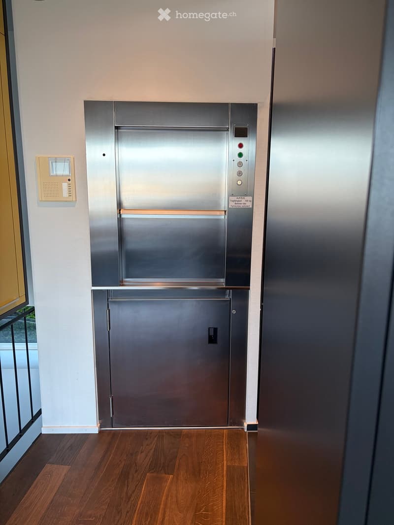 Modern hallway with wooden flooring, elevator door, and control panel on the wall.