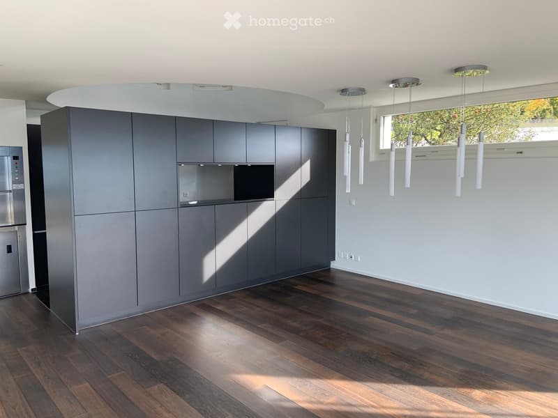 Modern kitchen with grey cabinets, wooden flooring, and a stainless steel refrigerator. Natural light streams in from a large window on the right.
