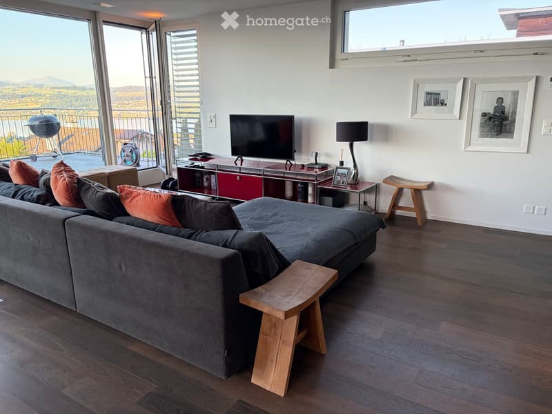 Modern living room with a grey sectional sofa, wooden floor, large windows offering a scenic view, a TV on a red and silver stand, and a floor lamp.