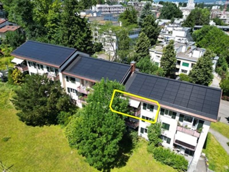 Aerial view of a multi-story residential building with solar panels on the roof, surrounded by greenery and neighboring structures.