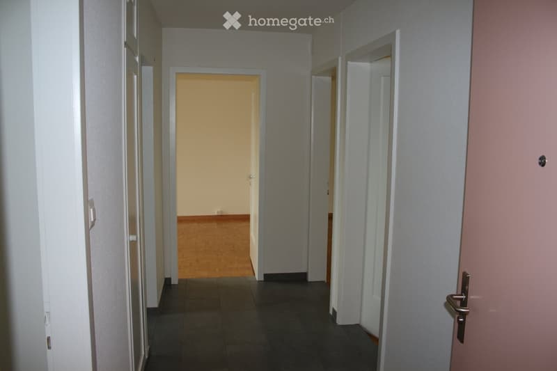 An empty hallway with tiled floor, wooden floor in one room, white walls, doors.