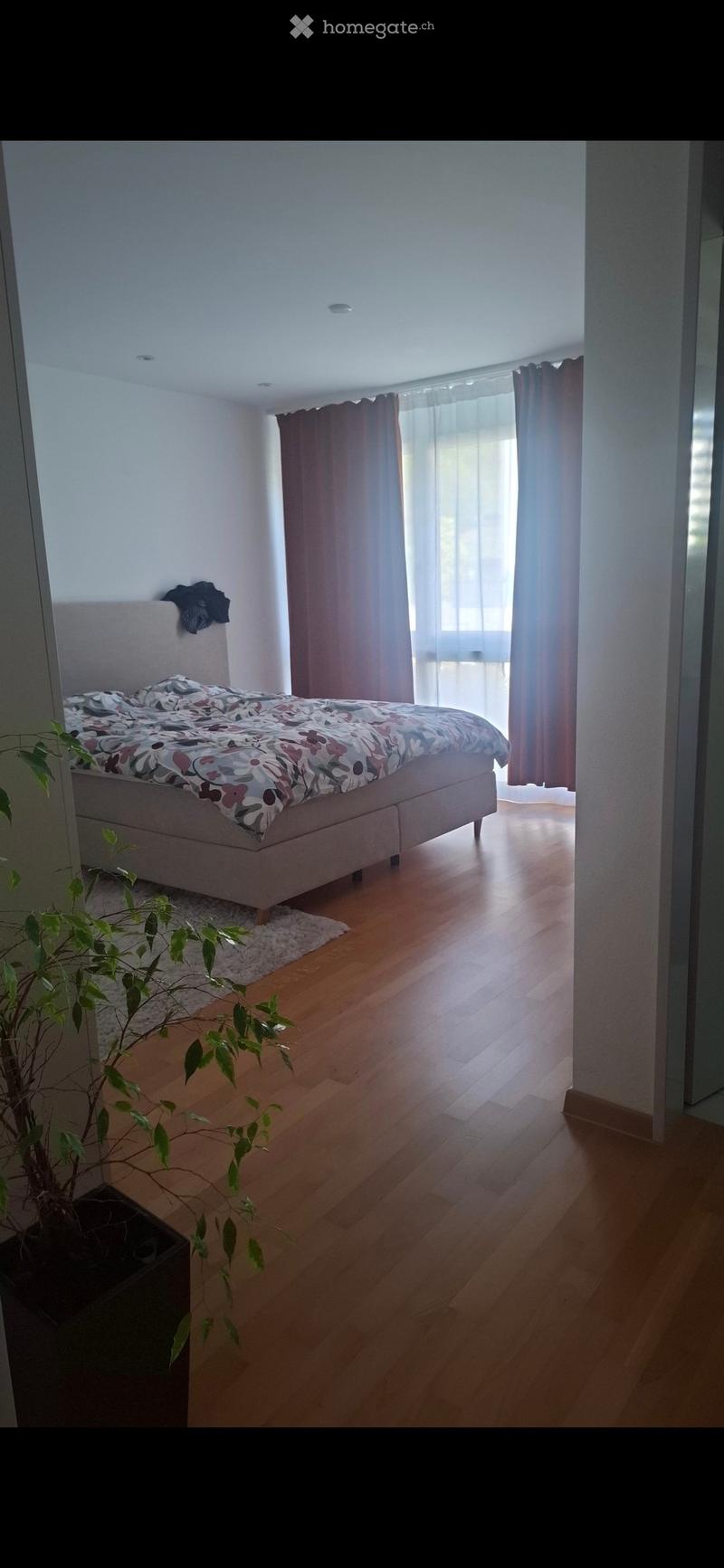 Modern bedroom with wooden flooring, large window with curtains, bed with floral duvet, small rug, and potted plant.
