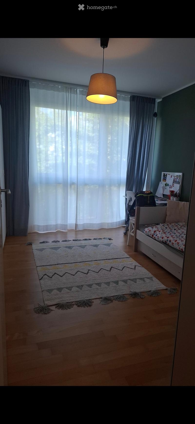 Bright bedroom with wooden flooring, large window with sheer curtains, a single bed with colorful bedding, a patterned rug, a desk with a chair, and a hanging lamp.
