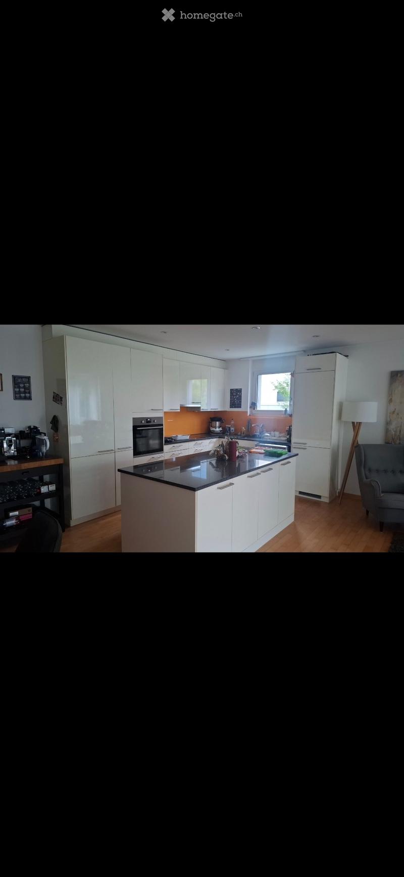 Modern kitchen with white cabinets, black countertop, and stainless steel appliances. Features a central island, a coffee maker on the counter, and a grey armchair in the corner.