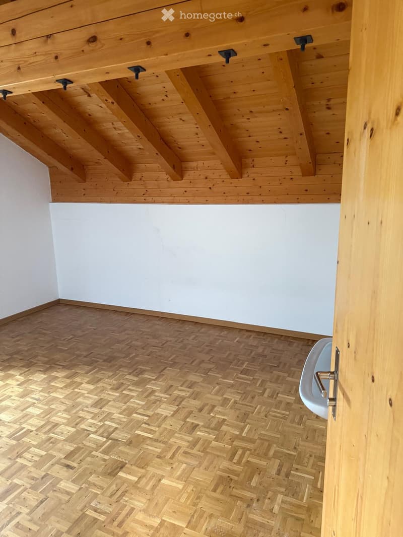 Empty room with wooden herringbone flooring and exposed wooden beams on the ceiling. White walls and a partially visible white bathtub in the adjacent space.