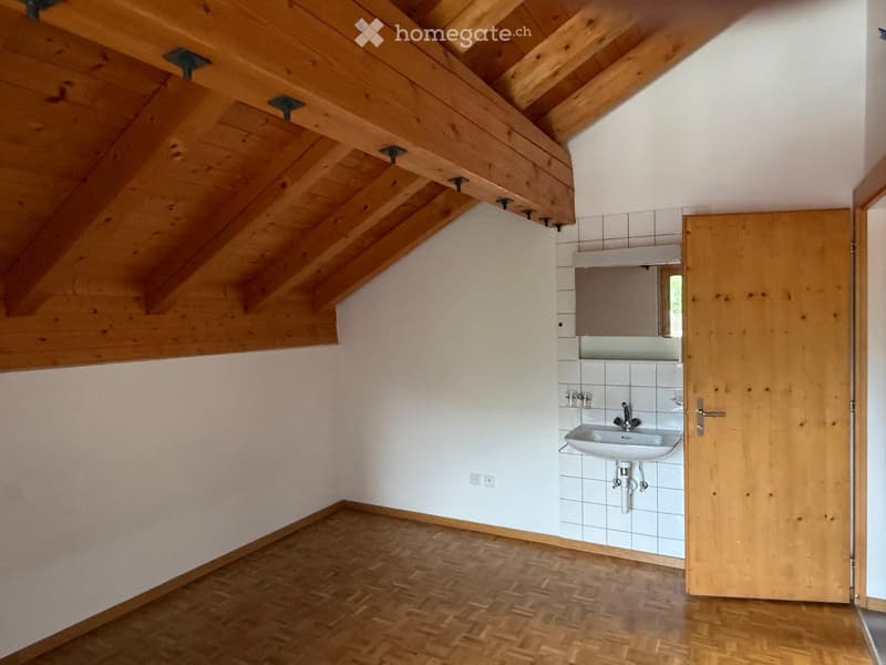An unfurnished attic room with wooden beams, white walls, and a small bathroom area featuring a sink and mirror.