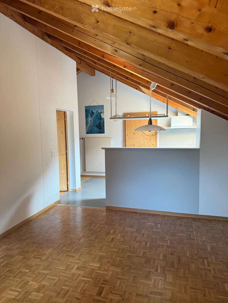 An unfurnished room with exposed wooden beams on the ceiling, wooden herringbone flooring, and a grey partition wall. The room features a hanging pendant light fixture and a doorway leading to another area.