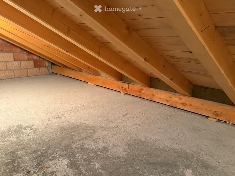 Empty attic space with wooden beams and concrete floor, exposed wooden framing, and brick wall visible at the edge.