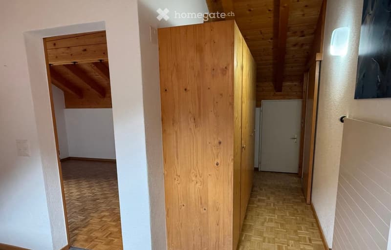 A narrow hallway with wooden flooring and white walls, featuring a tall wooden cabinet on one side and a partially visible room at the end.