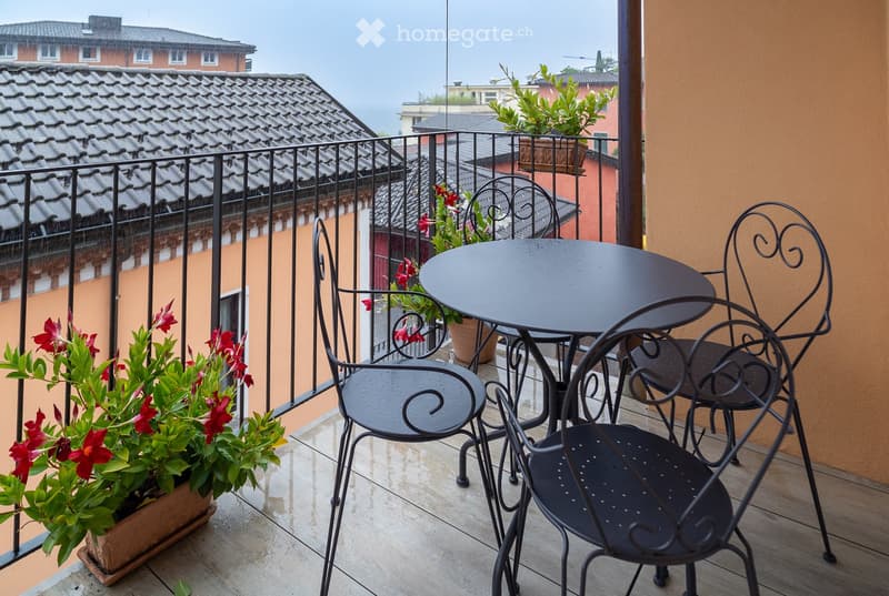 Balcony with a round black metal table, four matching chairs, and potted plants with red flowers.