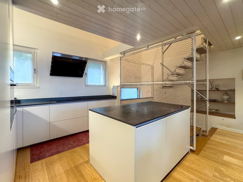 Modern kitchen with white cabinets, black countertops, wooden flooring, and a staircase leading to an upper level. A flat-screen screen is mounted on the wall.