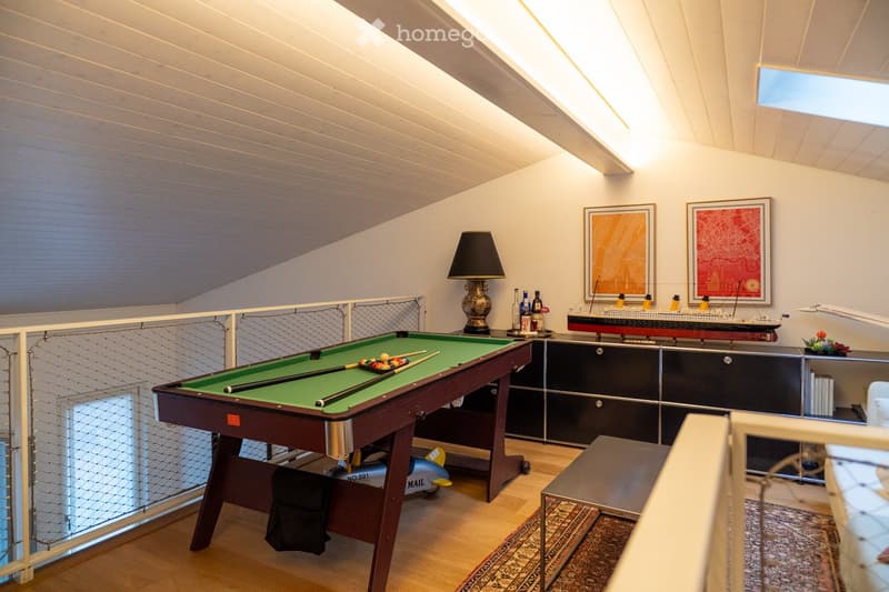 A well-lit room with a pool table, a table lamp, a model ship on a cabinet, framed artwork, and a small stool near the table.