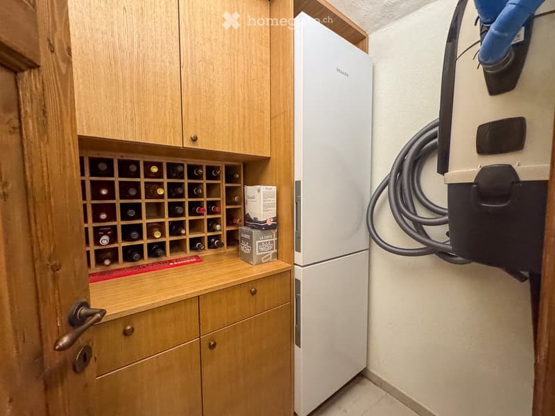 A small utility or storage room with wooden cabinets, a wine rack filled with bottles, a white refrigerator, and a few boxes on the counter.