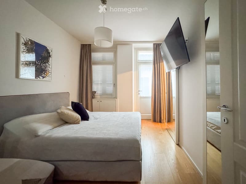 Modern bedroom with a large bed, pillows, headboard, pendant light, mirror, television, and cabinet. The room features wooden flooring and large windows with curtains.