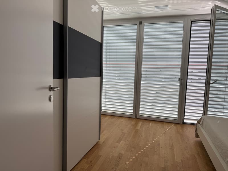 Modern bedroom with light wooden flooring, large sliding glass doors with blinds, and a single bed visible.