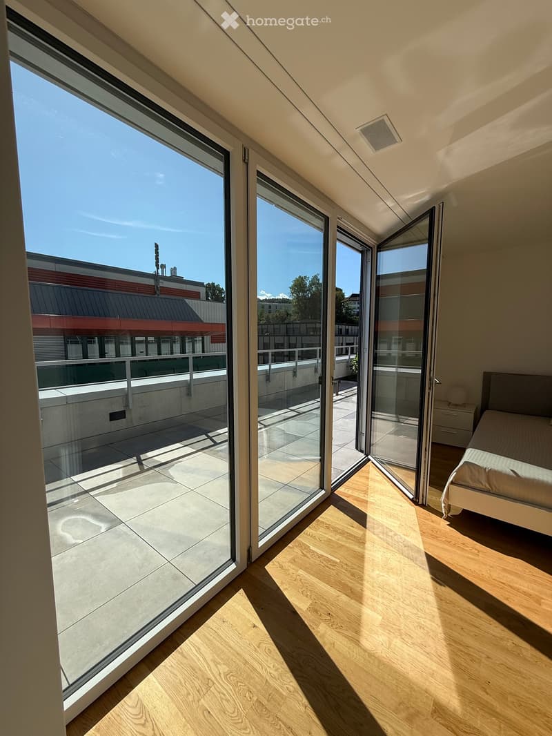 Bright bedroom with wooden flooring, large glass sliding doors leading to a balcony, and a neatly made bed.