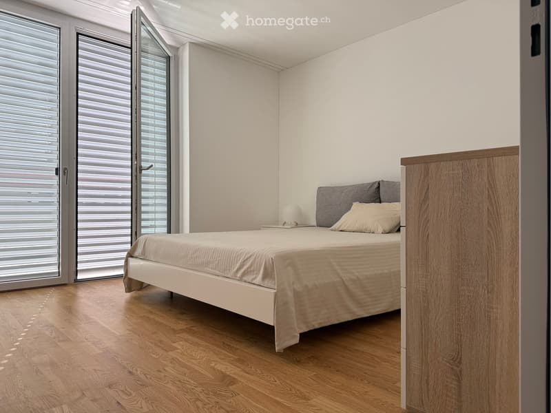 Modern bedroom with a white bed, grey pillows, a nightstand with a lamp, and large windows with blinds.