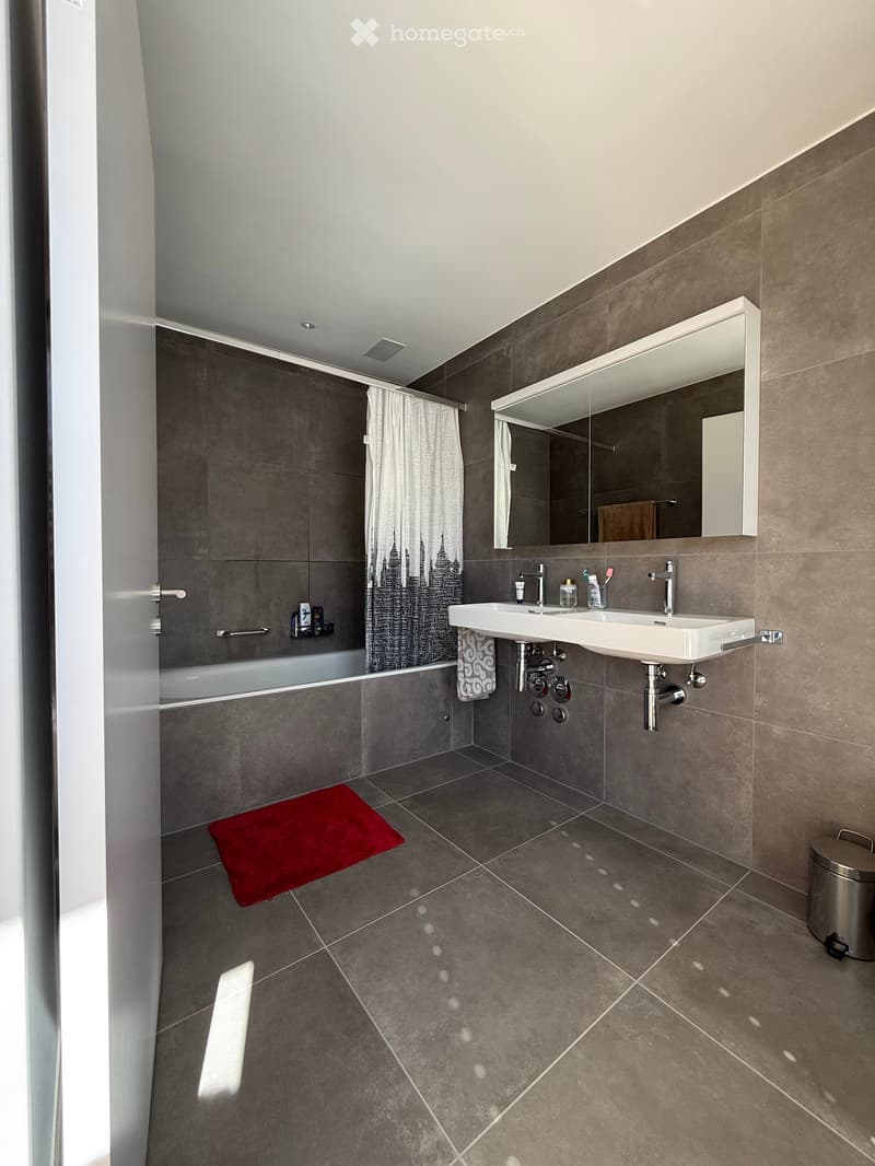 Modern bathroom with a double sink, large mirror, bathtub, and a red bathroom rug on the floor.