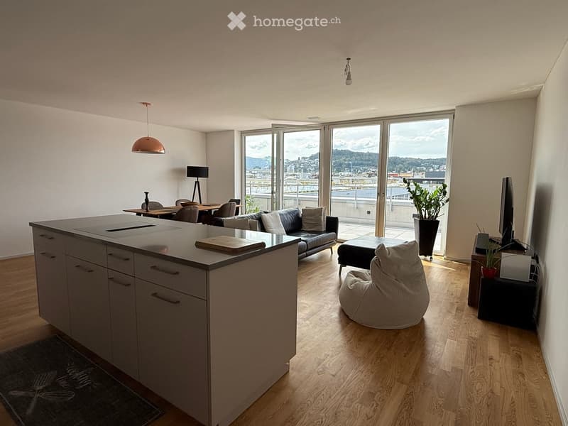 Modern open-plan living and dining area with a kitchen island, sofa, ottoman, beanie chair, dining table, chairs, pendant lamp, floor lamp, TV screen, and potted plants, with large windows offering a scenic view.