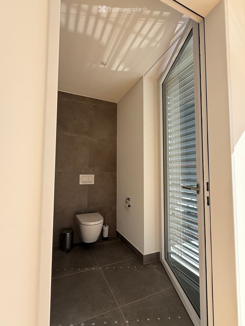 Modern bathroom with a toilet, dark tiled floor, grey walls, and a large window with blinds.