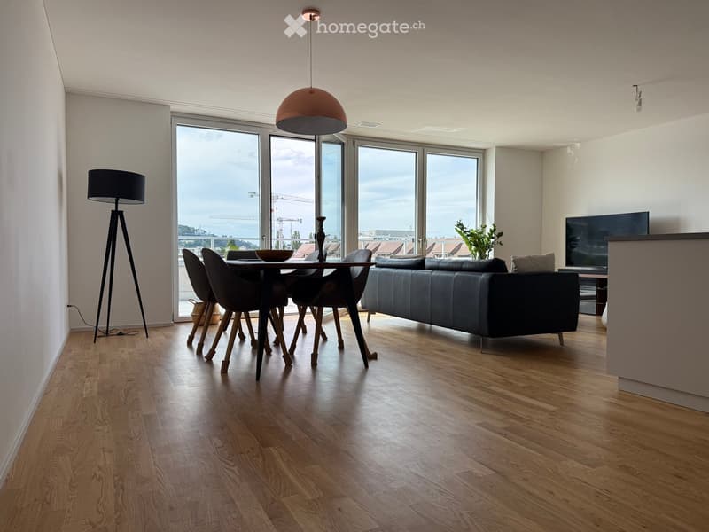 Modern living and dining area with hardwood floors, large windows offering a view, a dark blue sofa, a dining table with chairs, and a television on a stand.