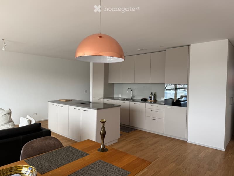 Modern open-plan living and kitchen area with white cabinets, grey countertops, a central island, a black sofa, a wooden dining table with chairs, and a large pendant light.