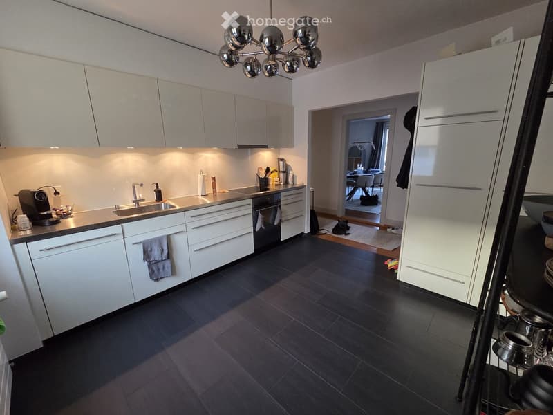 Modern kitchen with white cabinets, dark tiled floor, stainless steel sink, coffee maker, and a chandelier. A small dining area is visible through an open doorway.