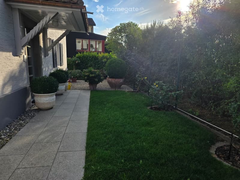 Well-maintained garden with a tiled pathway, neatly trimmed grass, potted plants, and a small fence. The area is bordered by a house with a covered entrance and a neighboring building visible in the background.