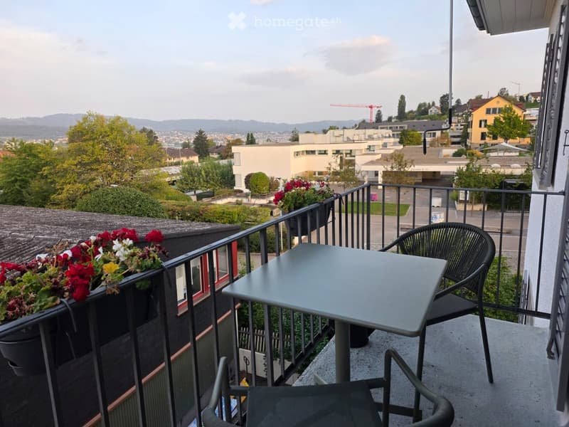 Balcony with a table and chairs, overlooking a cityscape with buildings and greenery.