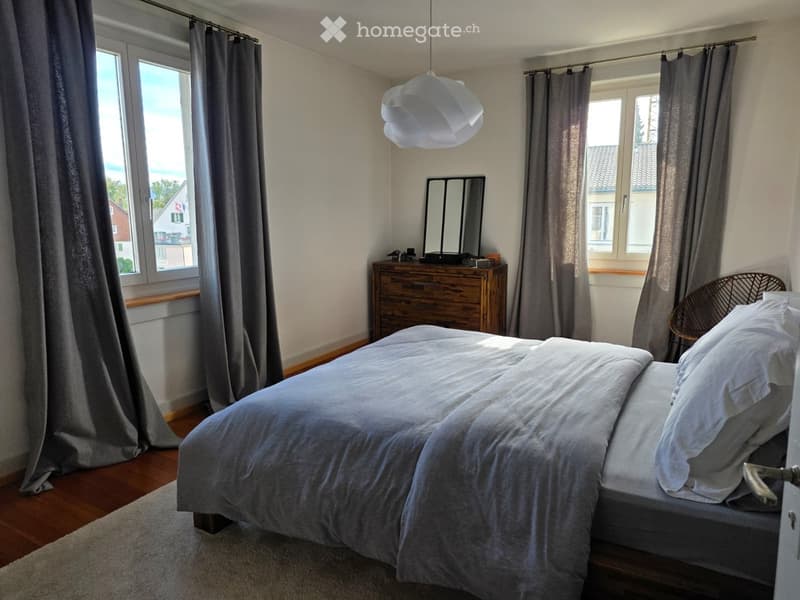 Cozy bedroom with wooden flooring, large windows, grey bedding, a wooden nightstand, a mirror, and a hanging lamp.