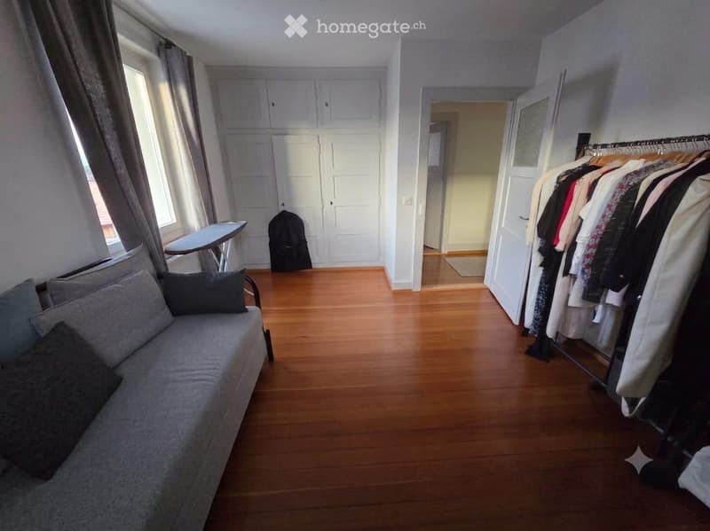 A furnished room with a grey sofa, pillows, an ironing board, a wardrobe, and a clothes rack with hanging garments.