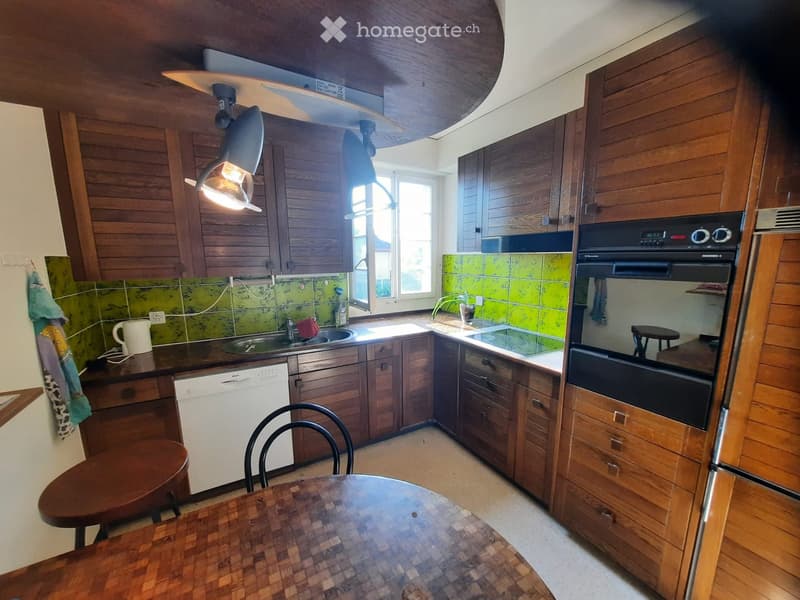 The kitchen features wooden cabinets, a central island, a sink, a dishwasher, an oven, and a small dining area with a table and chairs.