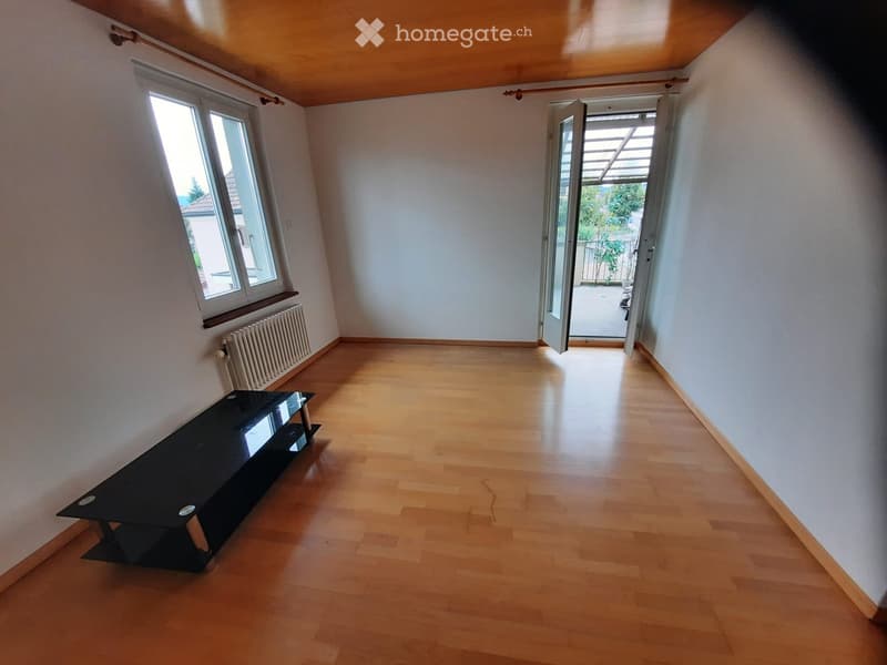 A small, unfurnished room with wooden flooring, white walls, and a single black table near the window. The room has an open doorway leading to a balcony or outdoor area.