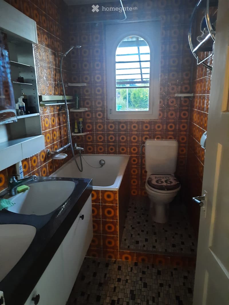 Bathroom with a sink, bathtub, toilet, and shelves. The walls are tiled with a circular pattern, and there is a window above the bathtub.