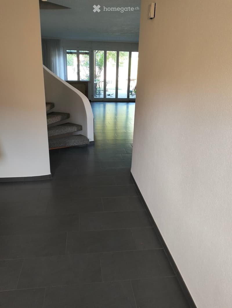Modern hallway with black tiled floor, white walls, curved staircase, large glass doors leading to outdoor area.