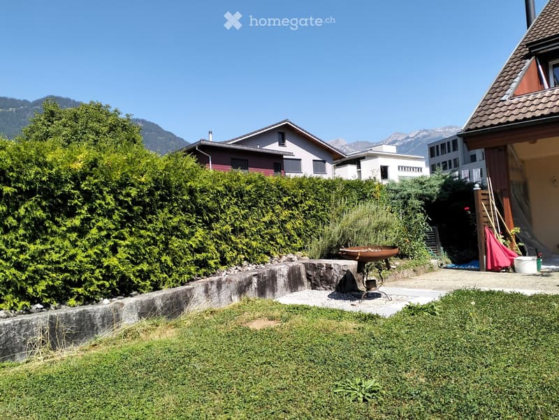 Garden area with a stone retaining wall, a metal fire bowl, and surrounding greenery, with residential houses and mountains in the background.