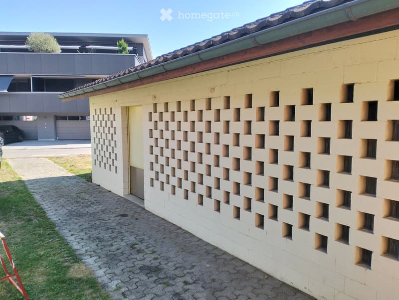 Exterior view of a modern building with a textured beige facade featuring a grid of square cutouts. The building has a tiled roof and a covered entrance area. A paved pathway leads to the entrance, with a grassy area on one side and a modern garage struct