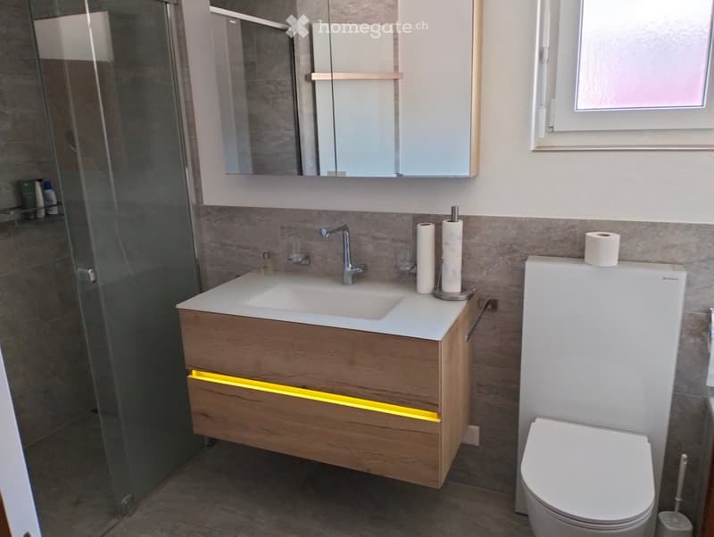 Modern bathroom with a wooden vanity, white sink, chrome faucet, toilet, and a glass-enclosed shower area.